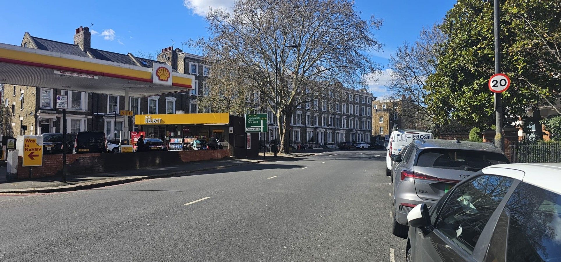 Une rue bordée de voitures garées, une station-service Shell et un panneau indiquant la limite de vitesse.
