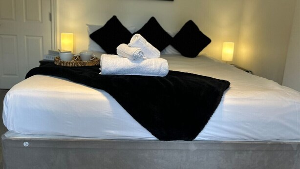 A neatly made bed with white linens, black pillows, and a black throw blanket. There are two bedside lamps and a framed picture on the wall.