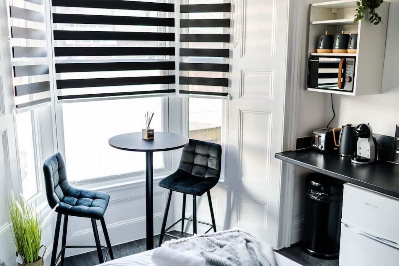 Et kompakt hotelværelse med en hjørneseng, to polstrede lænestole, et lille rundt bord, et tekøkken med kaffemaskine, brødrister og elkedel, samt et vindue med sort-hvide persienner.