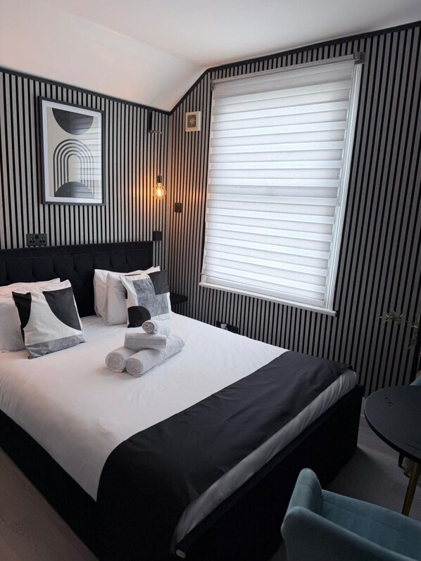 Habitación de hotel ordenada con una cama grande, una mesita de noche, una silla y una ventana con persianas.