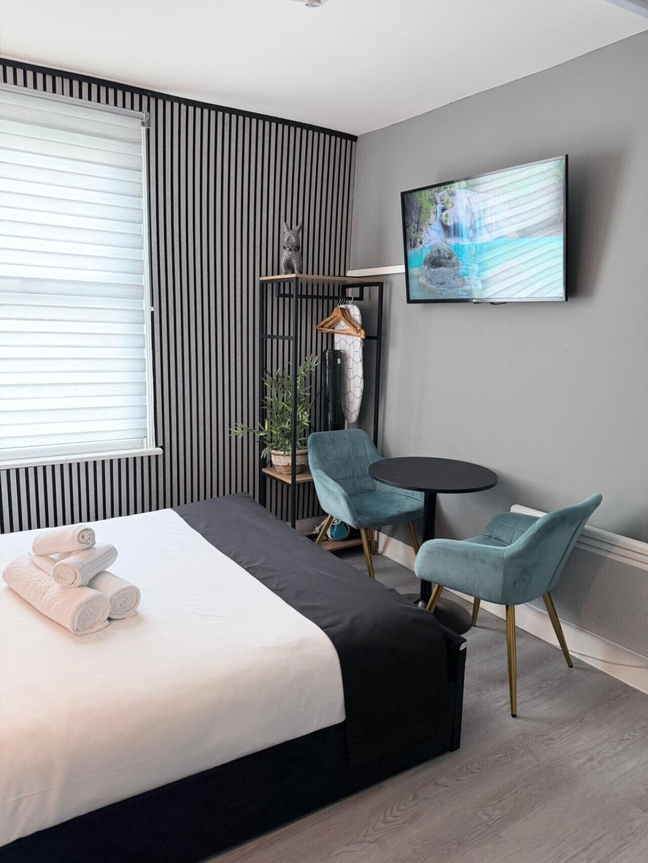 Ein modernes Hotelzimmer mit einem Bett, zwei Sesseln, einem kleinen Tisch, einem Fernseher und einem Fenster mit Jalousien.