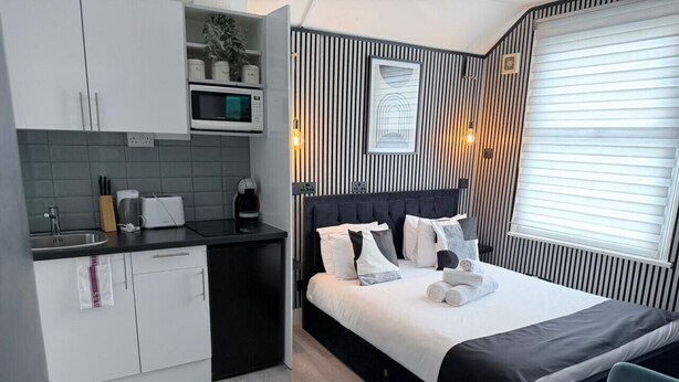 A compact hotel room with a kitchenette, a bed with multiple pillows, and a window with blinds.