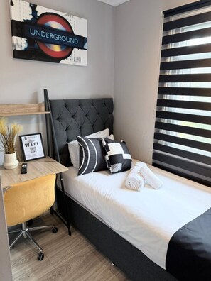 A hotel room with a bed, a desk with a chair, a potted plant, a framed picture, and a window with blinds.