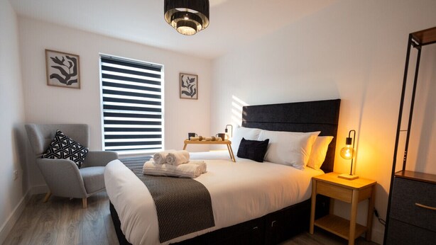 A modern bedroom with a large bed, a bedside table with a lamp, a chair, and artwork on the walls.