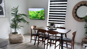 A modern living room with a grey sofa, a dining table with chairs, a TV on the wall, and a large window with blinds.
