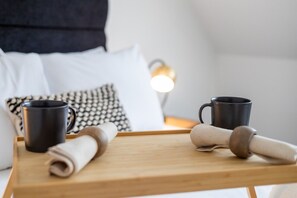 A wooden tray with two black mugs and a rolled-up beige towel on a bed with white pillows and a patterned pillow. - Market street 33A - 2 - second floor (Watford)