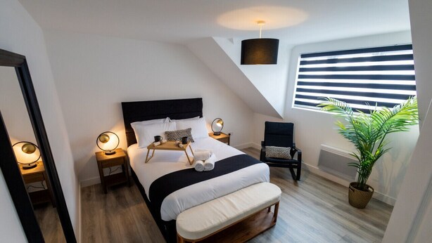 A modern bedroom with a large bed, bedside tables with lamps, a chair, a potted plant, and a window with blinds. - Market street 33A - 2 - second floor (Watford)
