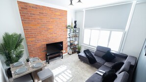 A living room with a brick wall, a grey sofa, a TV, and a dining area with a table set for four.