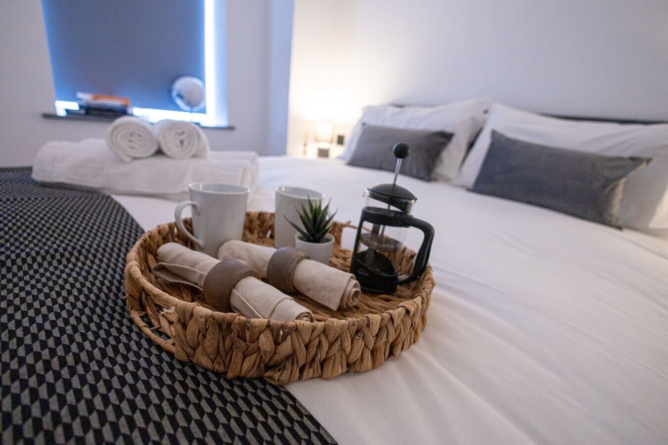 Un letto rifatto con cura, coperto da una coperta a quadri bianchi e neri, un comodino con una macchina da caffè, asciugamani arrotolati e una piccola pianta in vaso.