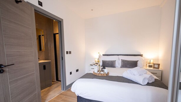 A modern hotel room with a large bed, bedside tables, a nightstand, and a bathroom visible through an open door. - Avenir Court Flat 5 (Finchley) (Greater London)