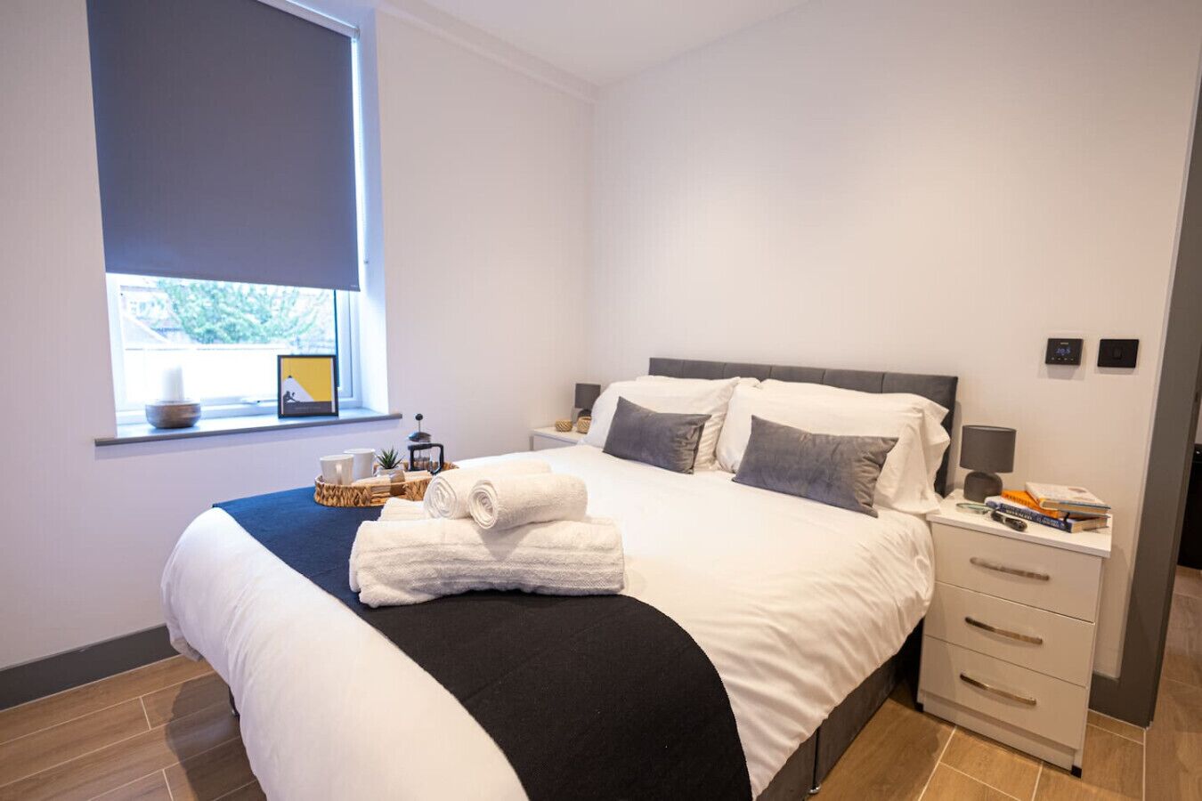 A modern hotel room with a large bed, a bedside table with a lamp, and a window with a view of greenery.