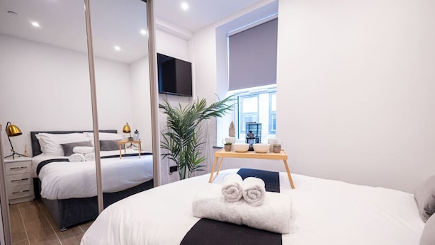 A modern hotel room with a large bed, a bedside table with a lamp, a TV mounted on the wall, and a window with a view of the city. - Avenir Court Flat 6 (Finchley) (Greater London)