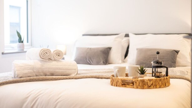 Una cama bien hecha con una colcha blanca, dos toallas enrolladas, una mesita de noche con una cafetera y una pequeña planta.