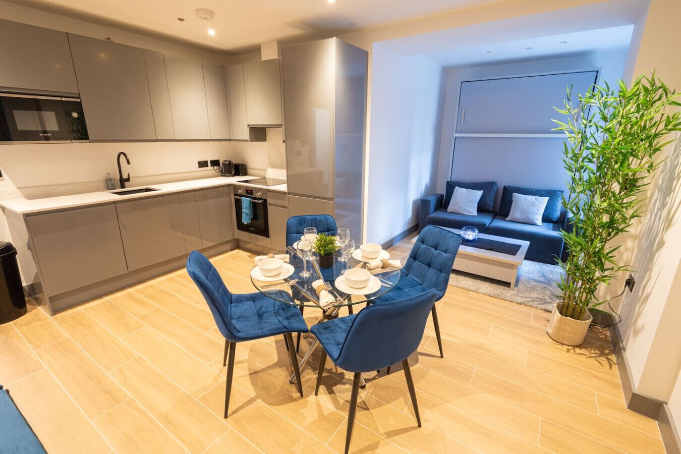 A modern kitchen with grey cabinets, a dining area with blue chairs, and a living room with a sofa and a potted plant.