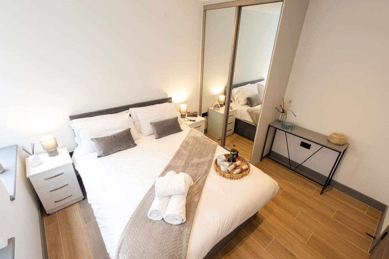 A modern hotel room with a large bed, bedside tables, a mirror, and a small side table with a lamp and decorative items.