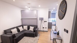 A modern living room with a sectional sofa, a floor lamp, a clock, and a small table with a plant. - Avenir Court Flat 15 (Finchley) (Greater London)