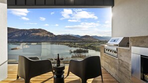 A balcony with a view of a lake and mountains, a grill, and two chairs.