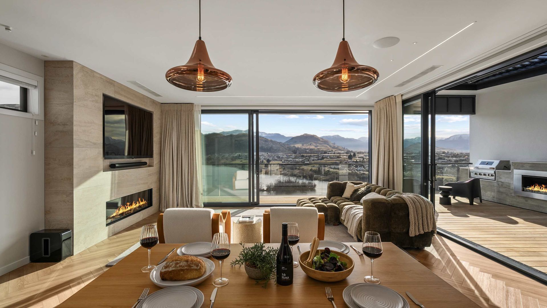 A modern dining area with a wooden table set for a meal, a fireplace, and a scenic view of a lake and mountains through large windows.