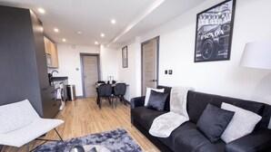 Una sala de estar moderna con un sofá de cuero negro, un sillón gris y una mesa de centro. Se observa un comedor con mesa y sillas, y una cocina al fondo.