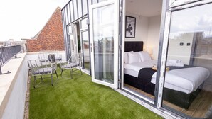 Una habitación de hotel moderna con puerta de vidrio que da a un balcón con césped artificial y sillas de metal.