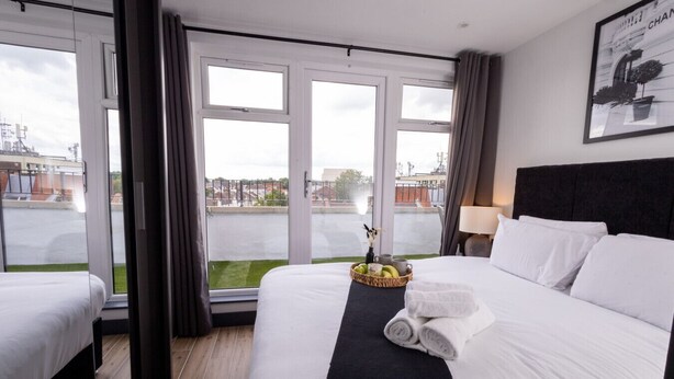 Habitación de hotel con una cama grande, una mesita con una cesta de frutas y vistas a la ciudad a través de amplios ventanales.