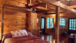 A cozy log cabin bedroom with a large bed, wooden ceiling beams, and a view of the outdoors through the windows. - Creekside B&B Cabin – Breakfast Included – Award-Winning Appalachian Farm (Irvine)