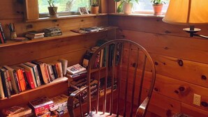 A cozy cabin interior with wooden walls, a rocking chair, a bookshelf, and a window with a view of trees. - Creekside B&B Cabin – Breakfast Included – Award-Winning Appalachian Farm (Irvine)