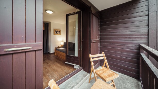 A wooden balcony with a wooden chair and a small table, a door leading to an interior room with a bed and a nightstand, and a framed picture on the wall.