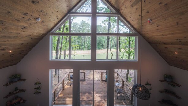 Amplia habitación en el ático con grandes ventanales que ofrecen vistas a un área boscosa y una terraza con mobiliario exterior.