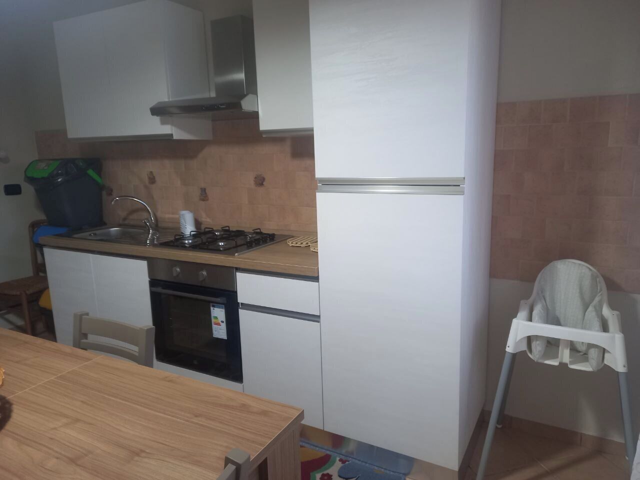 A compact kitchen with a stove, sink, and a white refrigerator. There is a wooden table and a white chair.