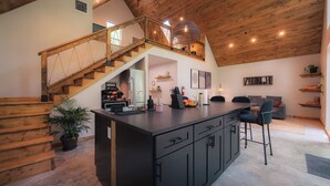 A modern kitchen with a central island, wooden staircase, and a lofted dining area. - #5 Woodpecker at Lost Forest Cabins (Richards)