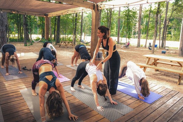 Un groupe de personnes pratiquant le yoga sur des tapis en bois, sous une structure également en bois, dans un parc.