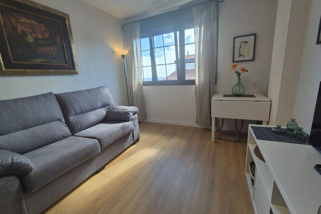 A living room with a grey sofa, a small white table with a vase, a painting on the wall, and a window with curtains.