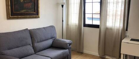 Une pièce avec un canapé gris, un tableau au mur, un bureau et une fenêtre avec des rideaux.