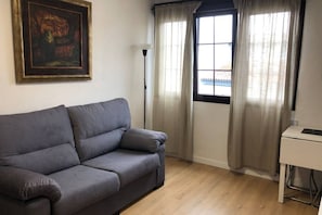 A room with a grey sofa, a painting on the wall, a desk, and a window with curtains.
