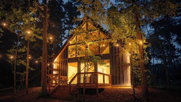 Uma cabana com deck de madeira e escadas que levam a porta de vidro, rodeada por árvores e luzes de fada.