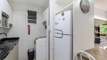 Una cocina con armarios blancos, una encimera negra, un refrigerador con imanes, un cubo de basura y un reloj en la pared.