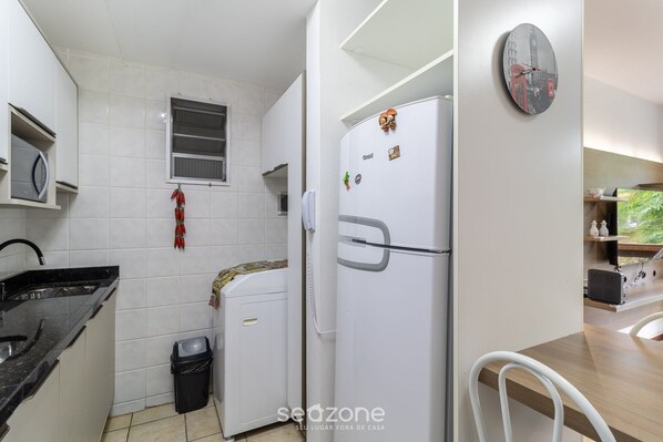 A kitchen with white cabinets, a black countertop, a refrigerator with magnets, a trash bin, and a clock on the wall.
