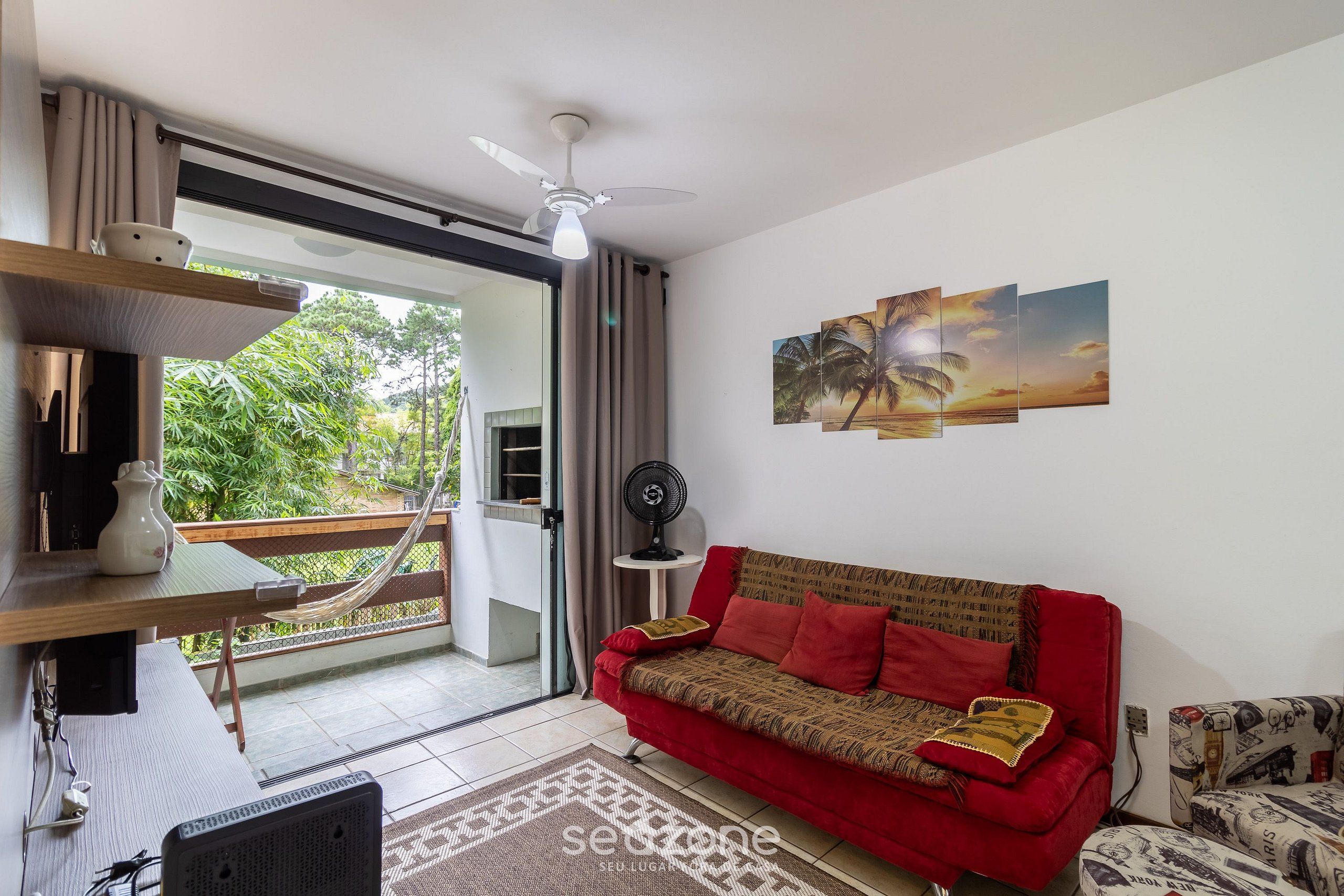 A living room with a red sofa, a ceiling fan, a wall-mounted TV, and a view of greenery outside.