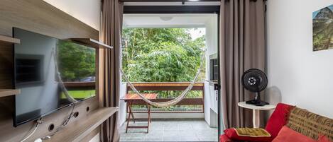 A living room with a flat-screen TV mounted on a wooden wall, a red sofa with patterned cushions, a small table with a fan, and a hammock on a balcony with a view of greenery.