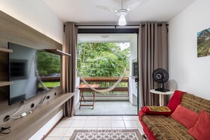 A living room with a flat-screen TV mounted on a wooden wall, a red sofa with patterned cushions, a small table with a fan, and a hammock on a balcony with a view of greenery. - 1 Bedroom Apt. With Barbecue Floripa-sc Abl0138 (Florianópolis)