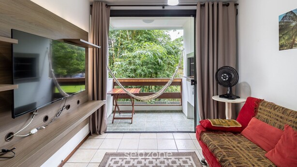 Una sala de estar con un televisor de pantalla plana montado en una pared de madera, un sofá rojo con cojines estampados, una mesita con un ventilador y una hamaca en un balcón con vistas al verdor.