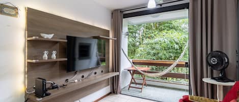 A modern living room with a flat-screen TV, wooden shelves, a hammock, a fan, and a balcony with greenery.