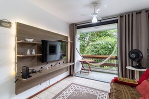 Una sala de estar moderna con un televisor de pantalla plana, estantes de madera, una hamaca, un ventilador y un balcón con plantas.