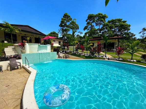 A swimming pool with clear blue water, surrounded by a landscaped garden with palm trees and a building in the background.