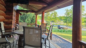 A covered outdoor seating area with wooden tables and chairs, a TV mounted on the wall, and a view of a road and greenery.