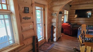 A log cabin interior with wooden walls, a wooden floor, a red leather sofa, a wooden dining table with chairs, and a flat-screen TV mounted on the wall.