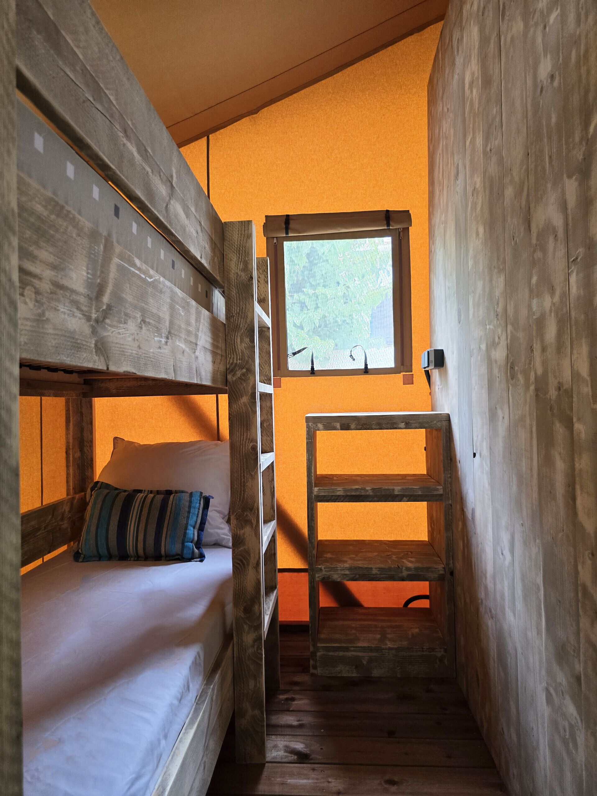 Una habitación pequeña y estrecha con una cama litera, una ventana que da a la vegetación y una silla de madera.
