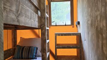 Una habitación pequeña y estrecha con una cama litera, una ventana que da a la vegetación y una silla de madera.