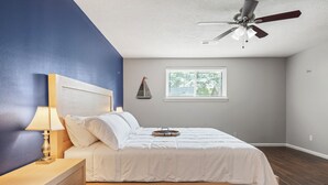 Een slaapkamer met een bed, nachtkastje, lamp, plafondventilator, raam en een decoratie aan de muur.